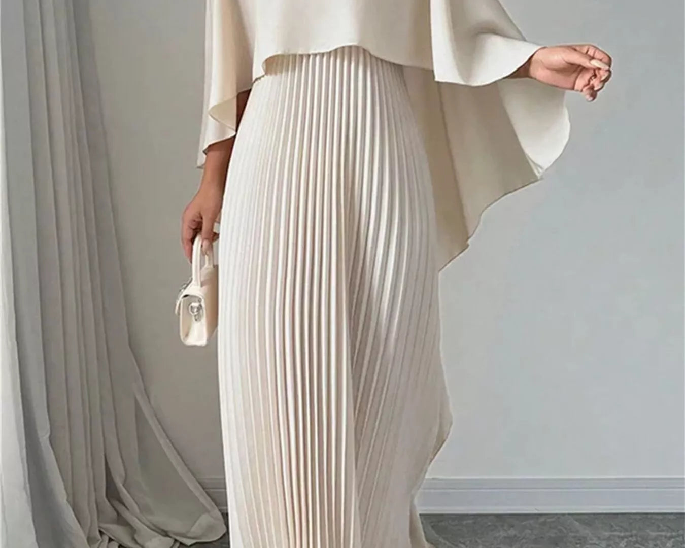 NEWYORKLISTED STYLE COLLECTION: Woman wearing a beige outfit with a draped top and pleated skirt against a plain background