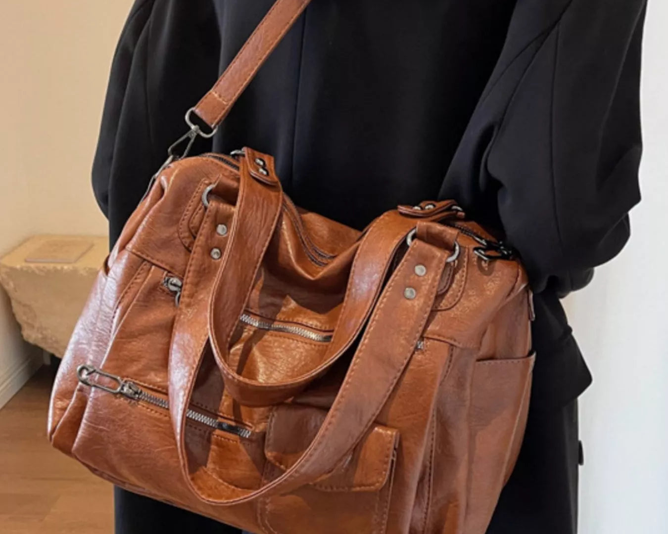 NEWYORKLISTED STYLE COLLECTION: Brown leather handbag held by a person wearing a black coat indoors.