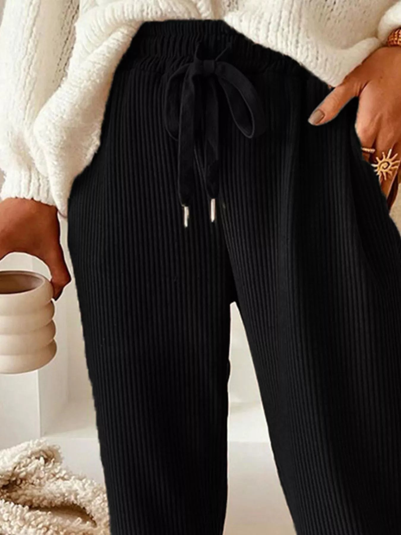 Person wearing black ribbed pants with a white sweater and beige blanket in the background