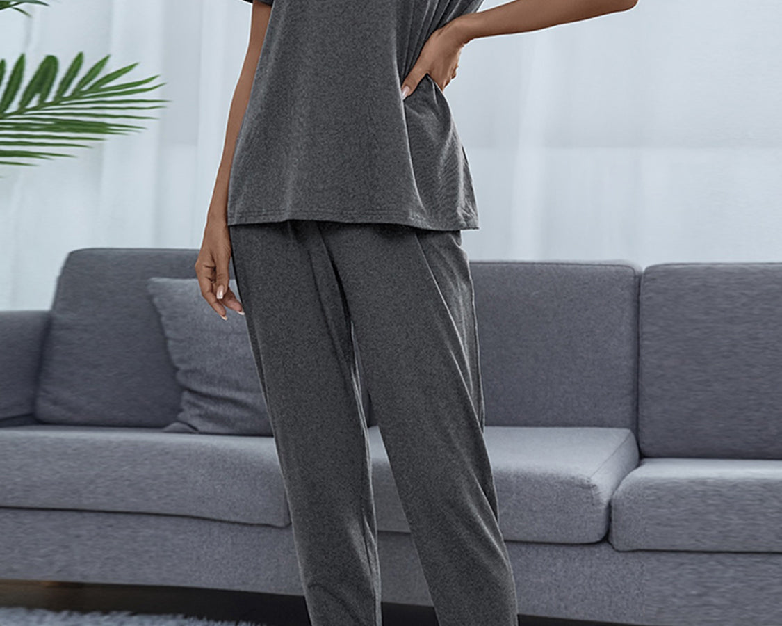 NEWYORKLISTED Intimate Apparel: Person wearing a gray pajama set standing in a living room with a gray sofa and white curtains.