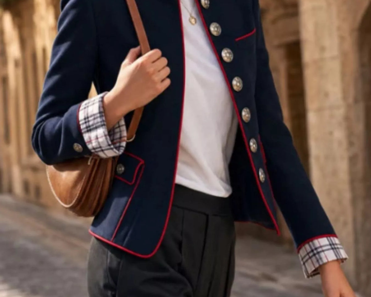 NEWYORKLISTED STYLE COLLECTION: A woman wearing a navy blue military-style blazer with red piping, silver buttons, and a plaid cuff, paired with a white top and dark trousers, carrying a brown shoulder bag in an outdoor stone alleyway.