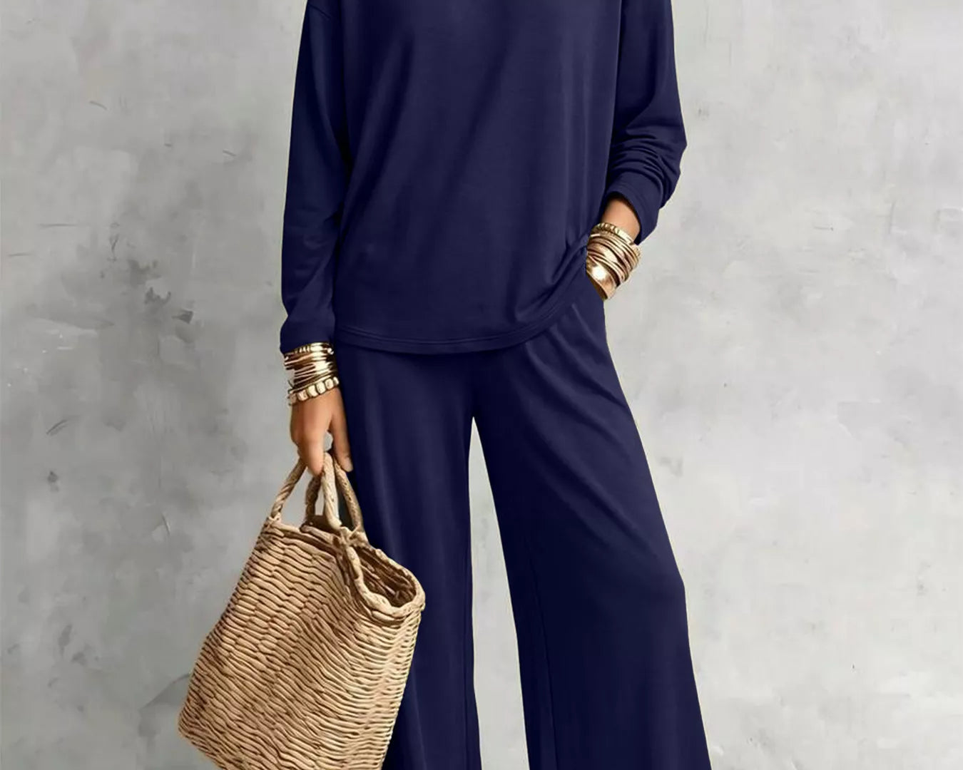 NEWYORKLISTED STYLE COLLECTION: A woman models a matching navy blue lounge set consisting of a long-sleeve top and wide-leg pants, styled with layered silver necklaces, gold bracelets, and a woven straw tote bag against a textured gray wall.