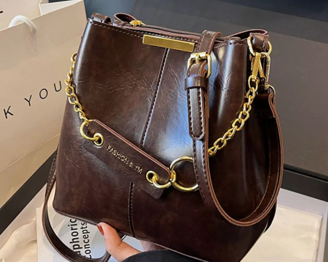 Brown leather handbag with gold chain details held by a person, on a white surface. New York Listed Style Collection