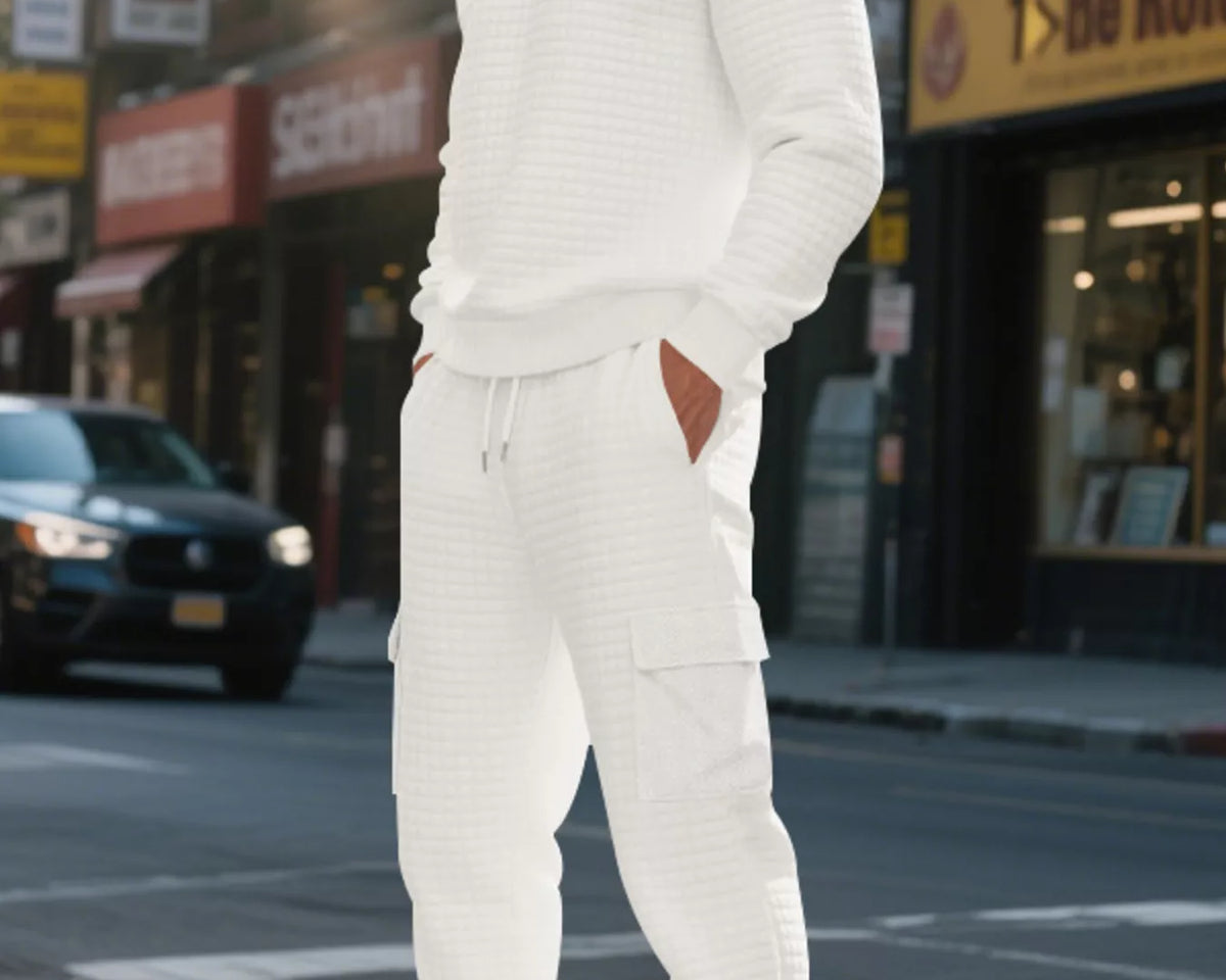 NEWYORKLISTED ATLAS MEN'S COLLECTION: A man wearing a matching white ribbed knit lounge set consisting of a long-sleeve crewneck sweater and jogger pants with cargo pockets, standing on a city street.