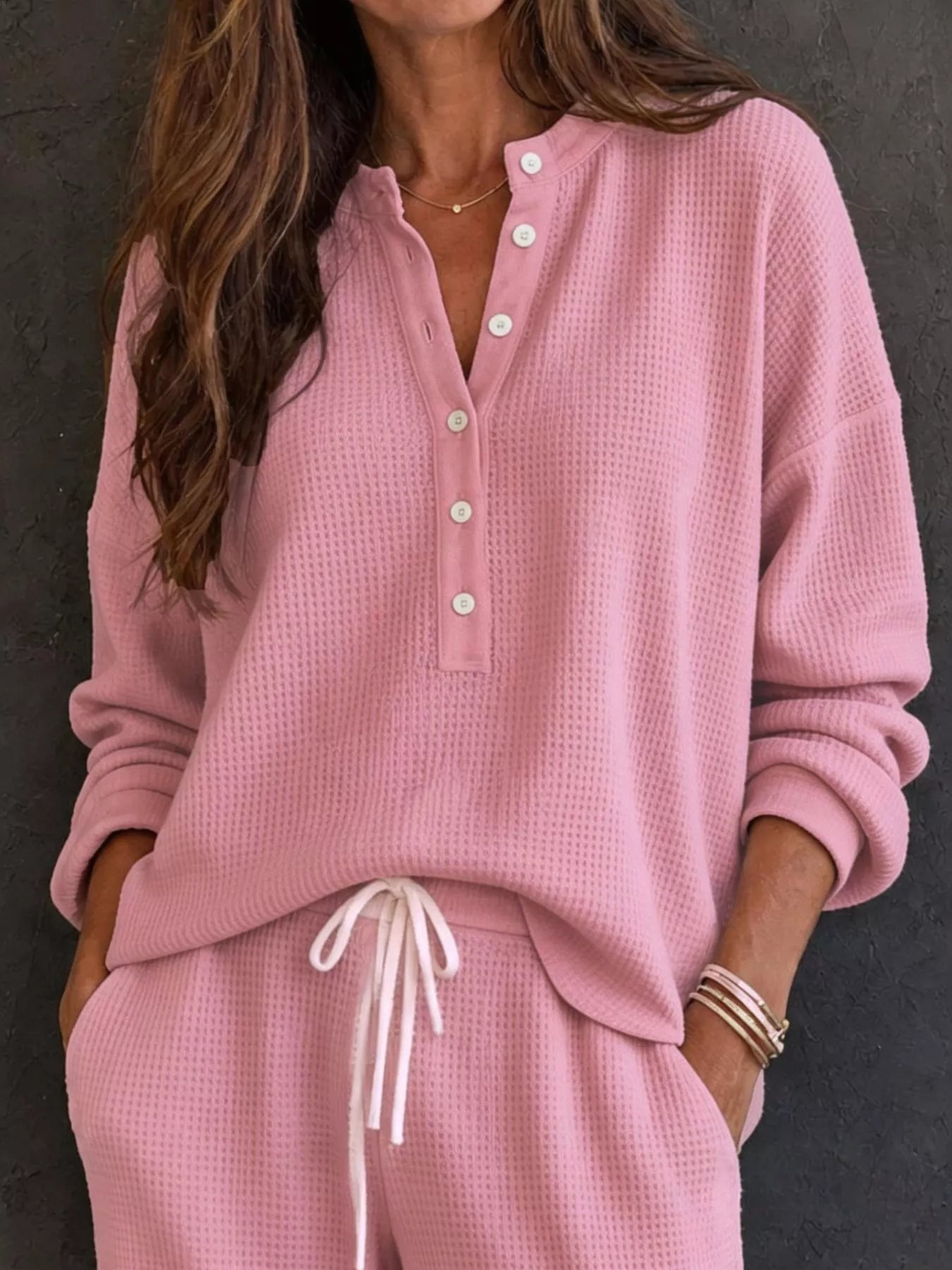 NEWYORKLISTED STYLE & COMFORT COLLECTION: A woman wearing a matching pink waffle-knit lounge set with a henley top and drawstring pants, photographed against a dark textured wall.