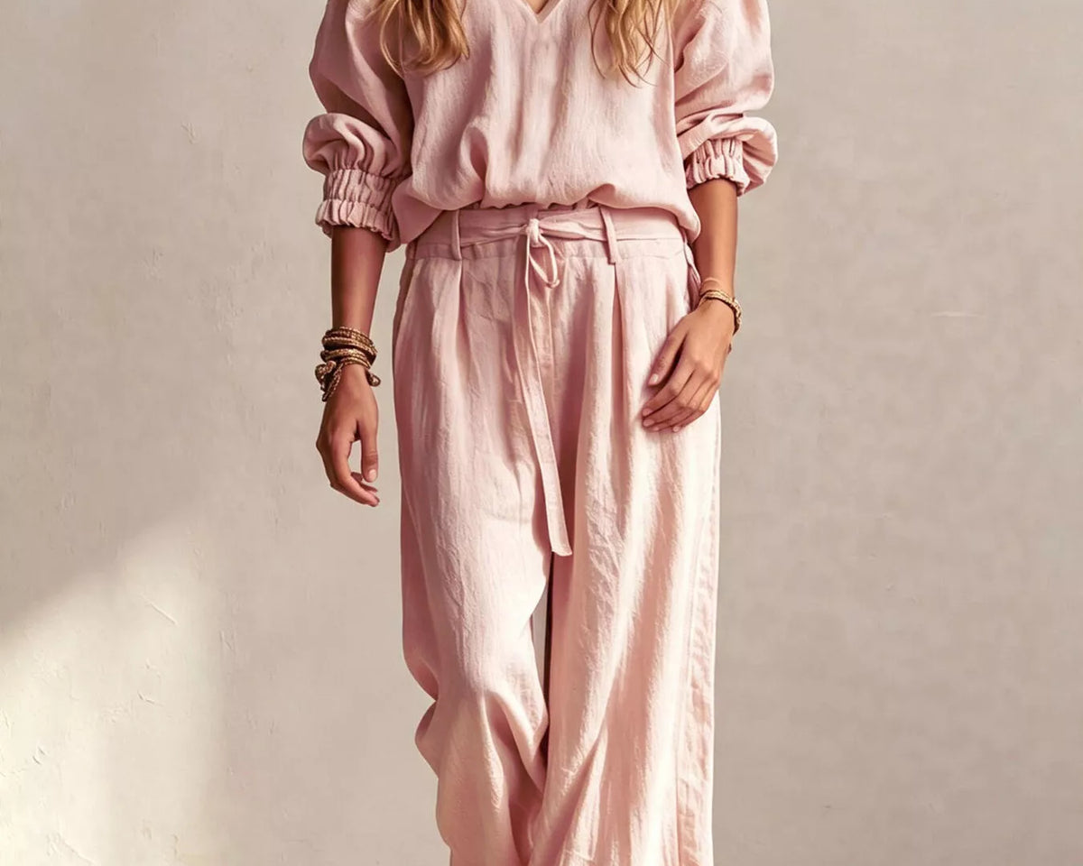 NEWYORKLISTED STYLE COLLECTION: A woman modeling a light pink two-piece linen set consisting of a long-sleeve V-neck top and matching wide-leg trousers, styled with layered necklaces and bracelets against a textured beige wall.