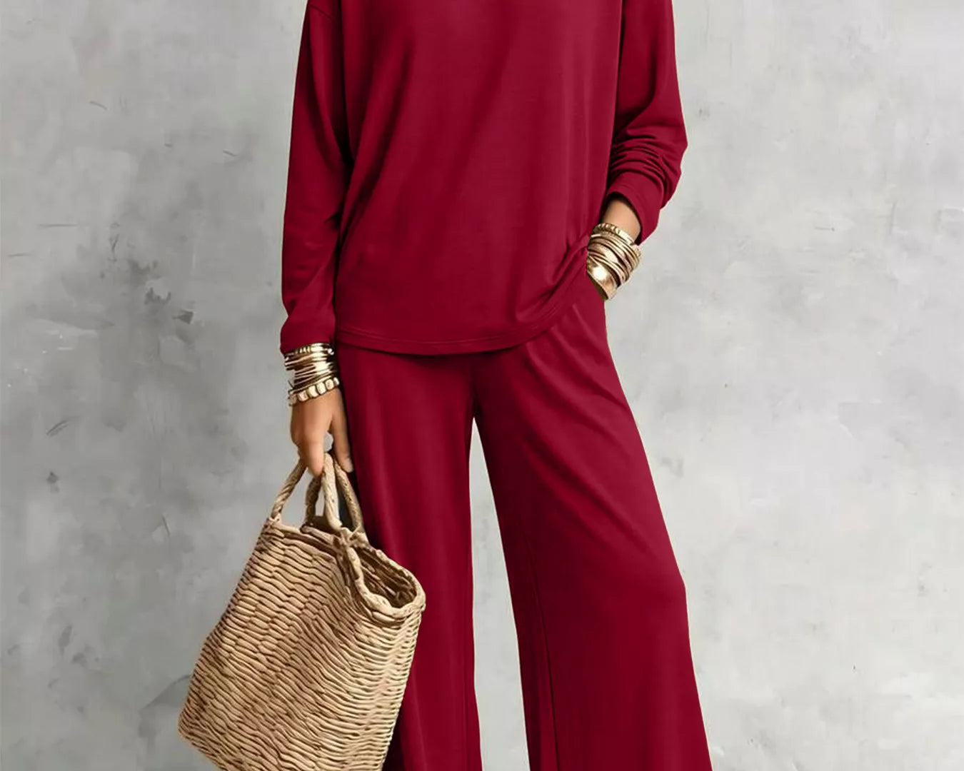 NEWYORKLISTED STYLE COLLECTION: A woman models a matching burgundy long-sleeve top and wide-leg pants set, styled with layered gold necklaces, bracelets, and a straw tote bag against a textured gray wall.