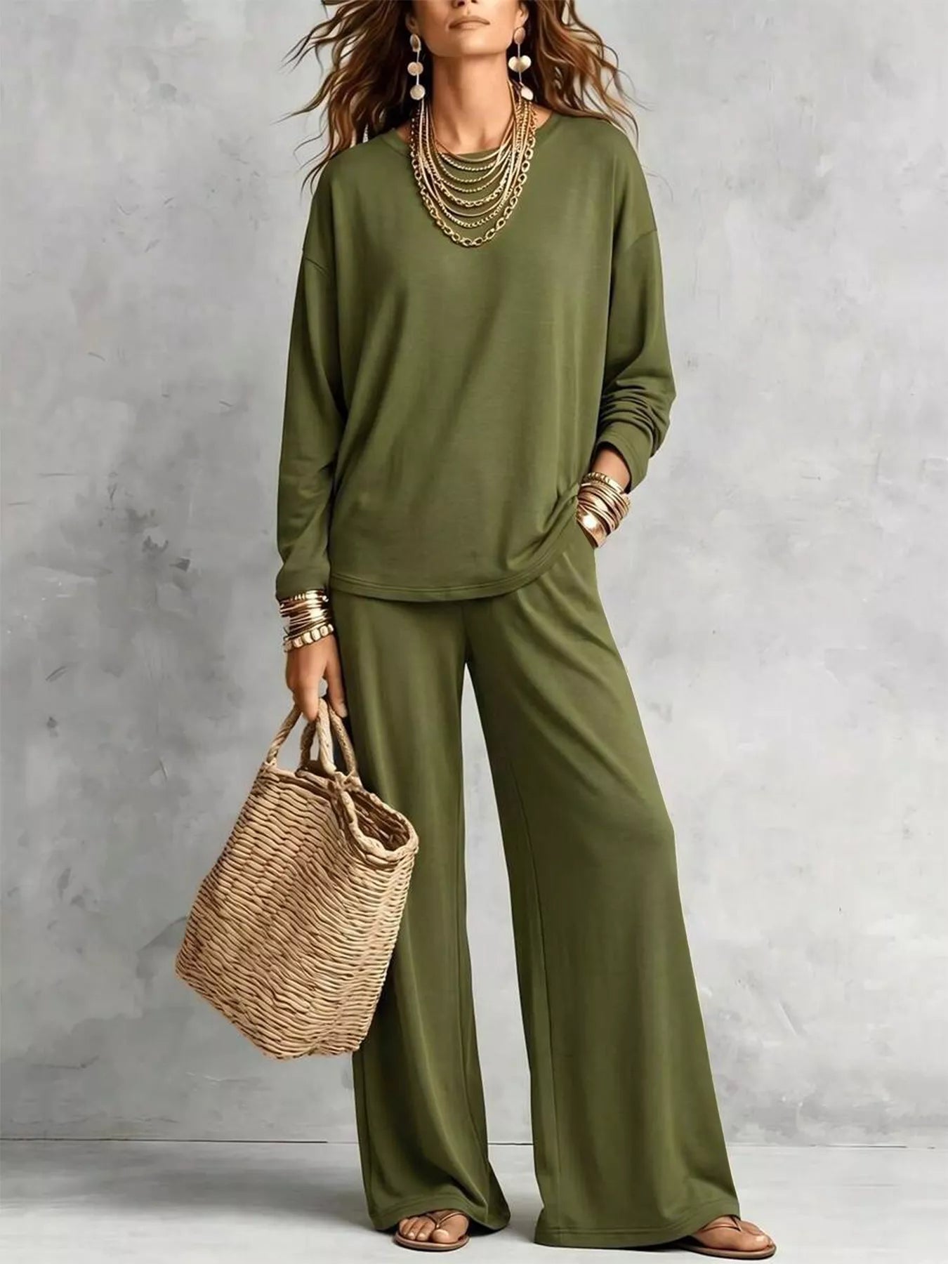 NEWYORKLISTED STYLE COLLECTION: A woman models an olive green two-piece lounge set consisting of a long-sleeve top and wide-leg pants, styled with layered gold necklaces, bracelets, and a straw tote bag against a textured gray wall.