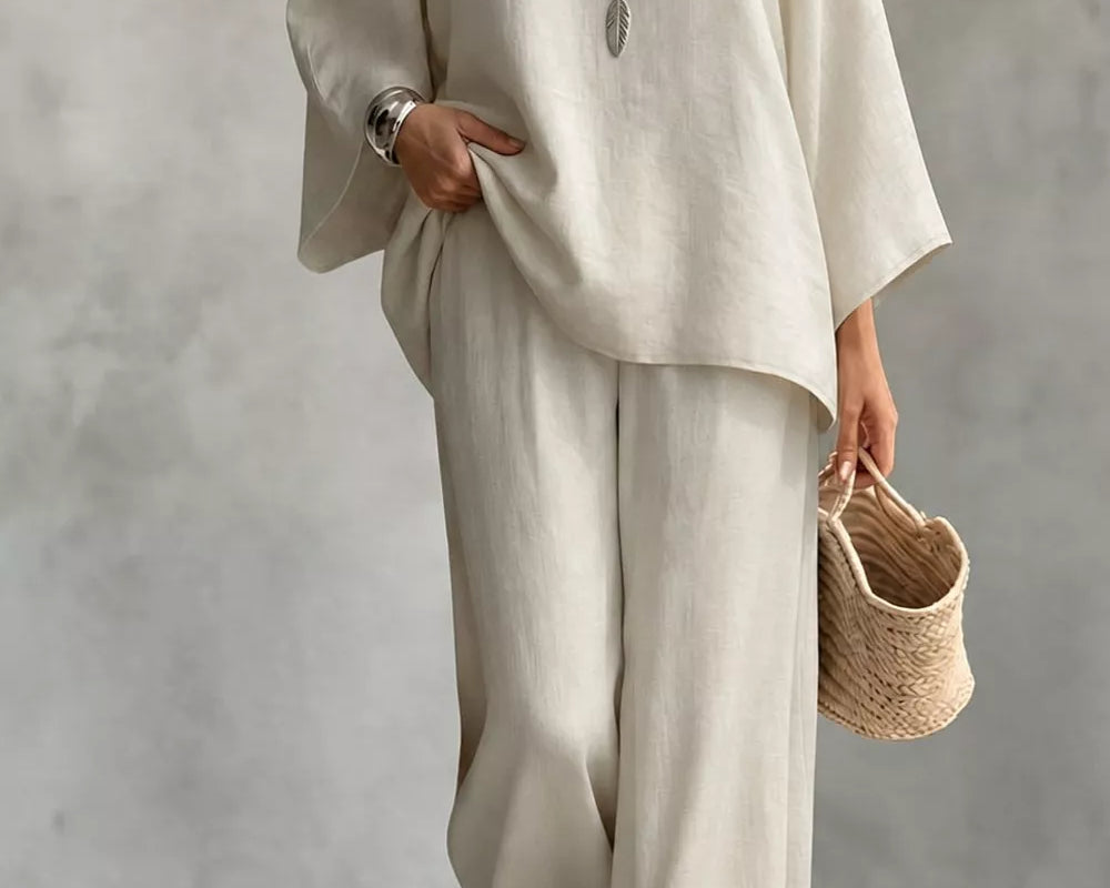 NEWYORKLISTED STYLE COLLECTION: A woman wearing a loose, two-piece linen outfit in a warm off-white or cream color, consisting of a wide-sleeve top and matching wide-leg pants. She is accessorized with layered silver necklaces, a silver watch, and holds a small woven straw handbag against a textured gray studio background.