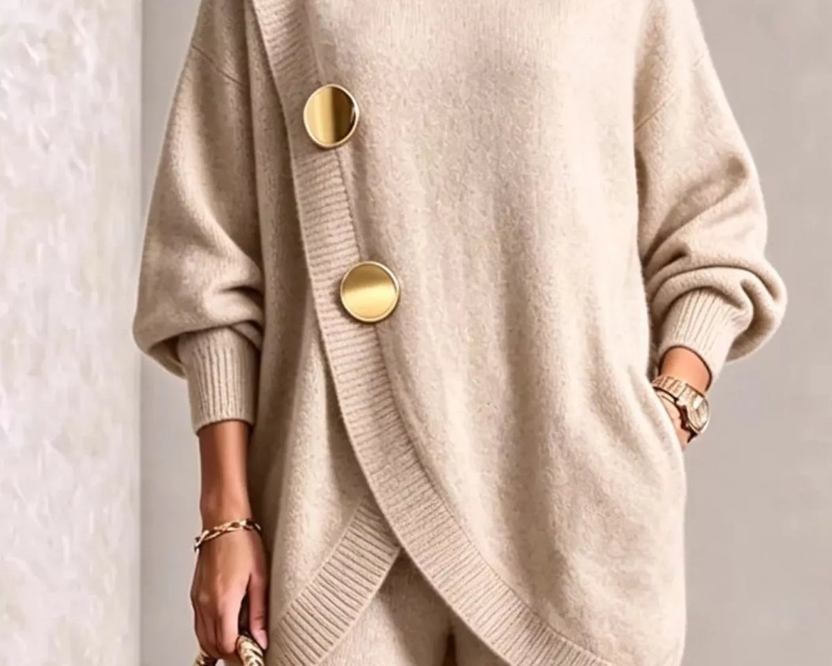 NEWYORKLISTED STYLE COLLECTION: A woman wearing a beige oversized turtleneck sweater with large gold-tone decorative buttons and matching beige pants, holding a small gold handbag against a light textured wall.
