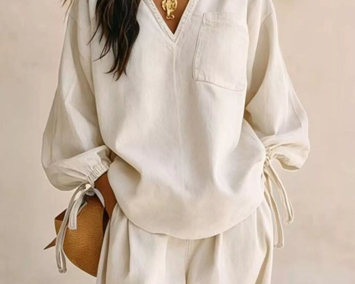 NEWYORKLISTED STYLE COLLECTION: A woman wearing a cream-colored linen two-piece set consisting of a V-neck long-sleeve top and matching wide-leg pants, styled with gold jewelry and a small brown woven bag against a neutral beige background.