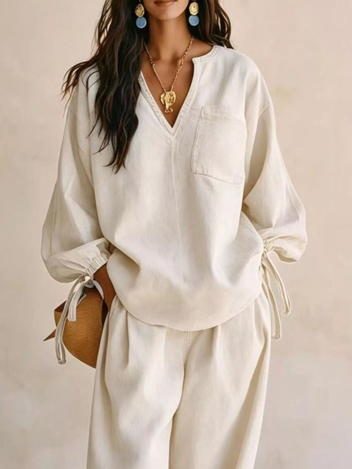 NEWYORKLISTED STYLE COLLECTION: A woman wearing a cream-colored linen two-piece set consisting of a V-neck long-sleeve top and matching wide-leg pants, styled with gold jewelry and a small brown woven bag against a neutral beige background.