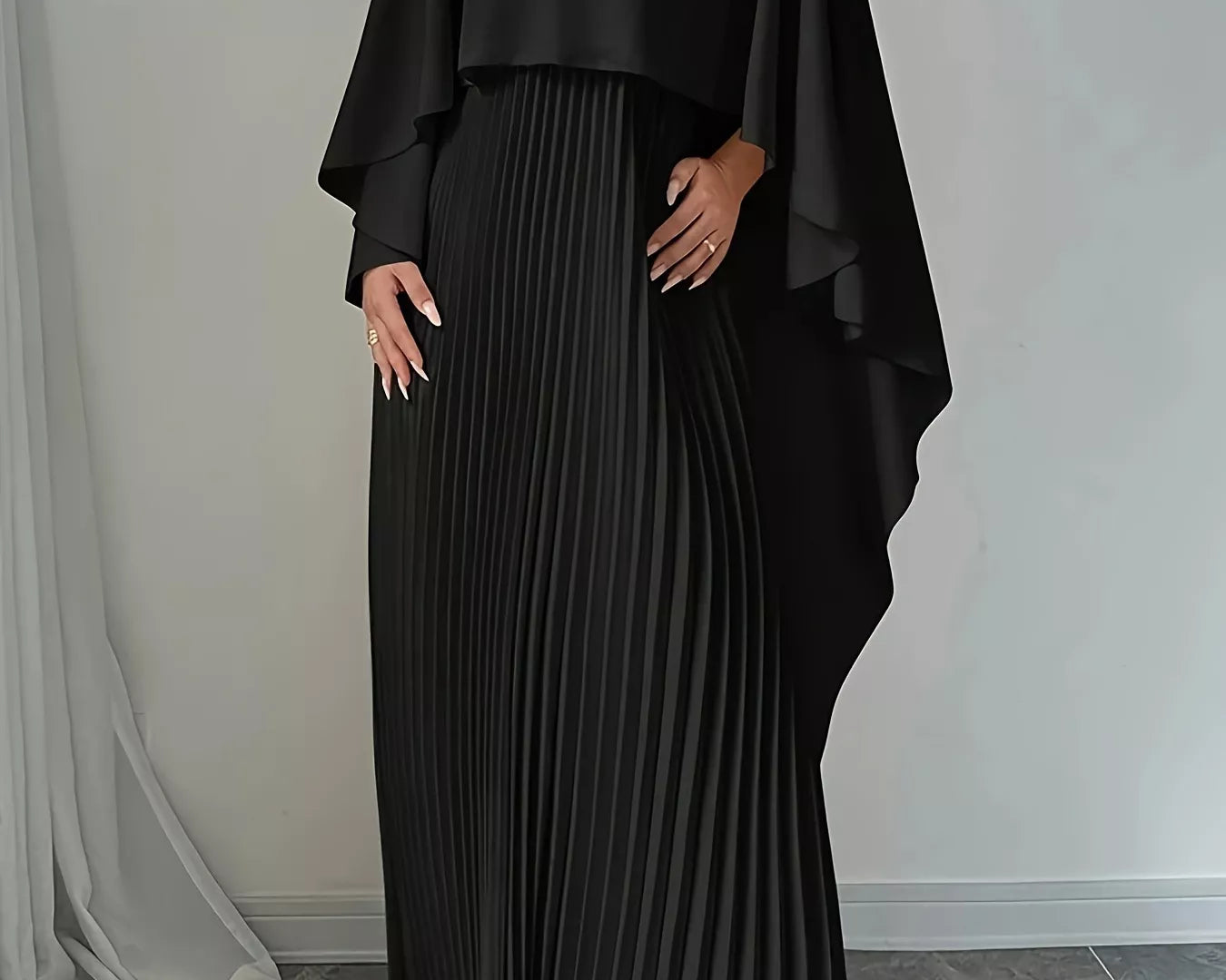 NEWYORKLISTED STYLE COLLECTION: Woman wearing a black pleated dress with a cape-like top against a neutral background