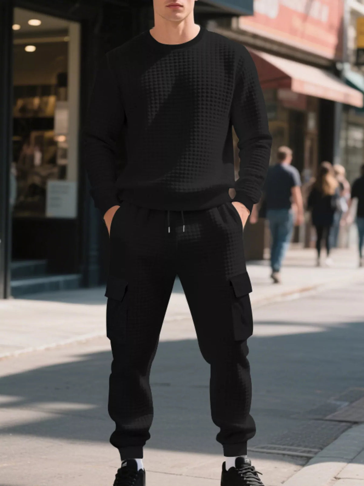 NEWYORKLISTED ATLAS MEN'S COLLECTION: A man wearing a black textured crewneck sweatshirt and matching black cargo jogger pants stands on a city sidewalk with blurred storefronts and pedestrians in the background.