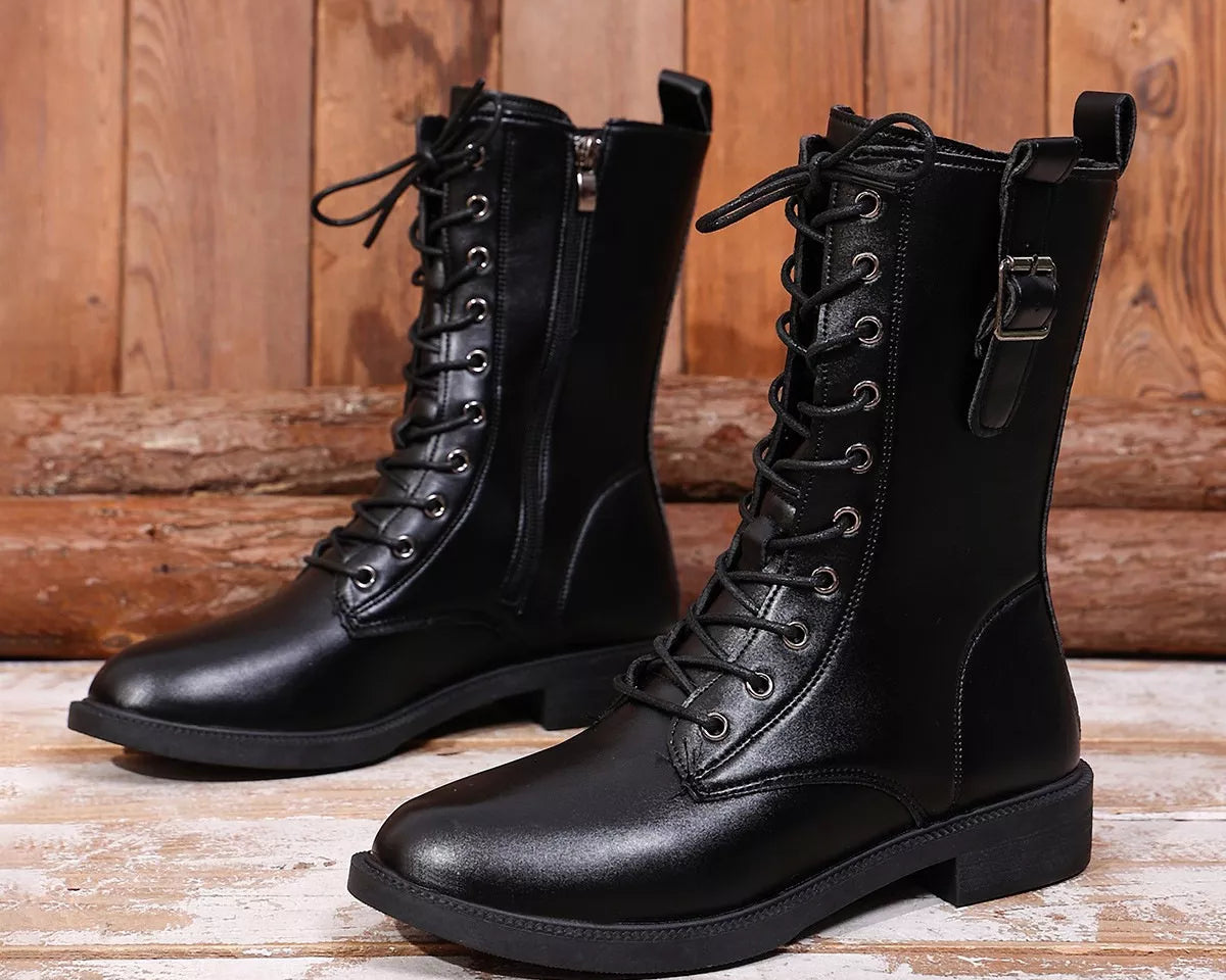 NEWYORKLISTED SHOE COLLECTION: A pair of black leather lace-up ankle boots with side zippers and buckle details, displayed on a rustic wooden plank floor against a wooden wall background.