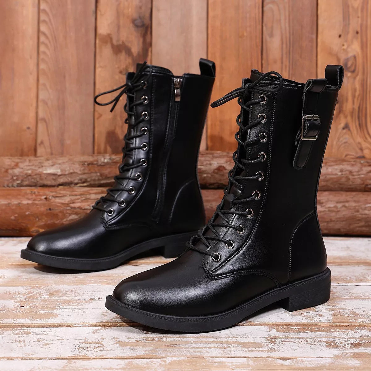 NEWYORKLISTED SHOE COLLECTION: A pair of black leather lace-up ankle boots with side zippers and buckle details, displayed on a rustic wooden plank floor against a wooden wall background.