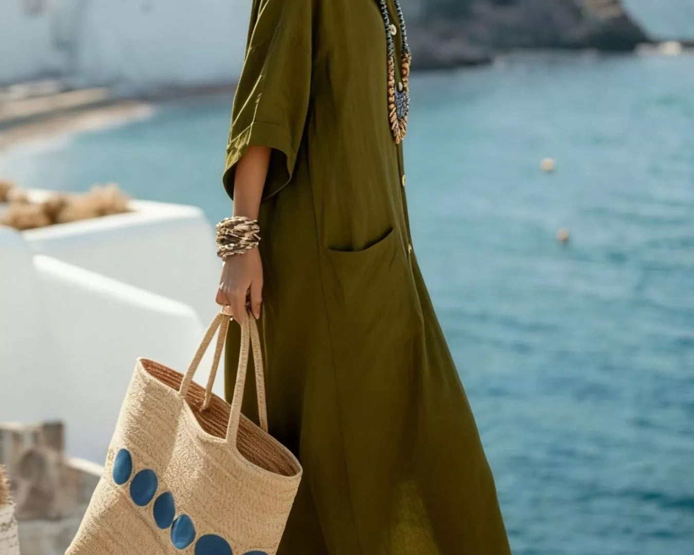 NEWYORKLISTED STYLE COLLECTION: Woman in a green dress holding a straw bag with blue accents, standing by the ocean.