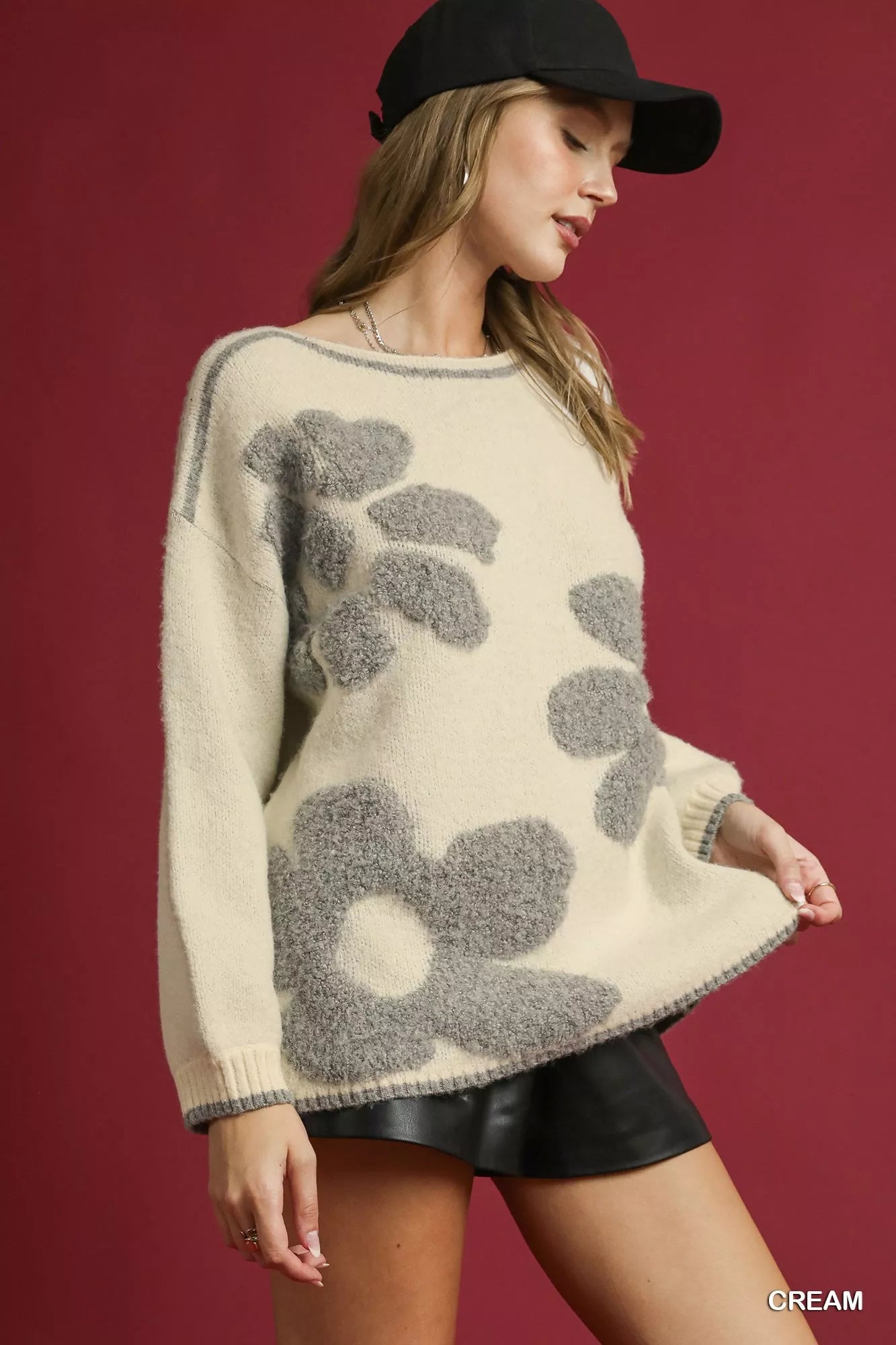 NEWYORKLISTED STYLE COLLECTION: Woman wearing a cream sweater with gray floral patterns and a black cap against a red background