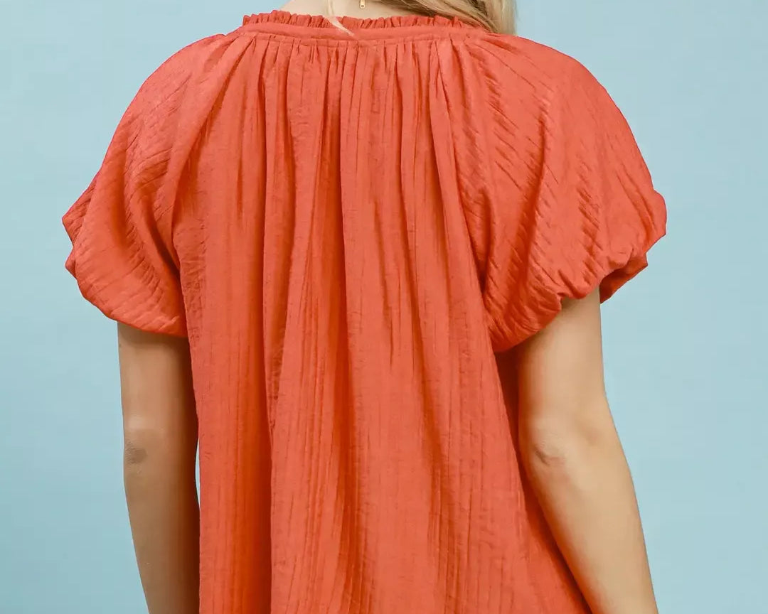 NEWYORKLISTED STYLE COLLECTION: Woman wearing a coral blouse with short sleeves against a light blue background