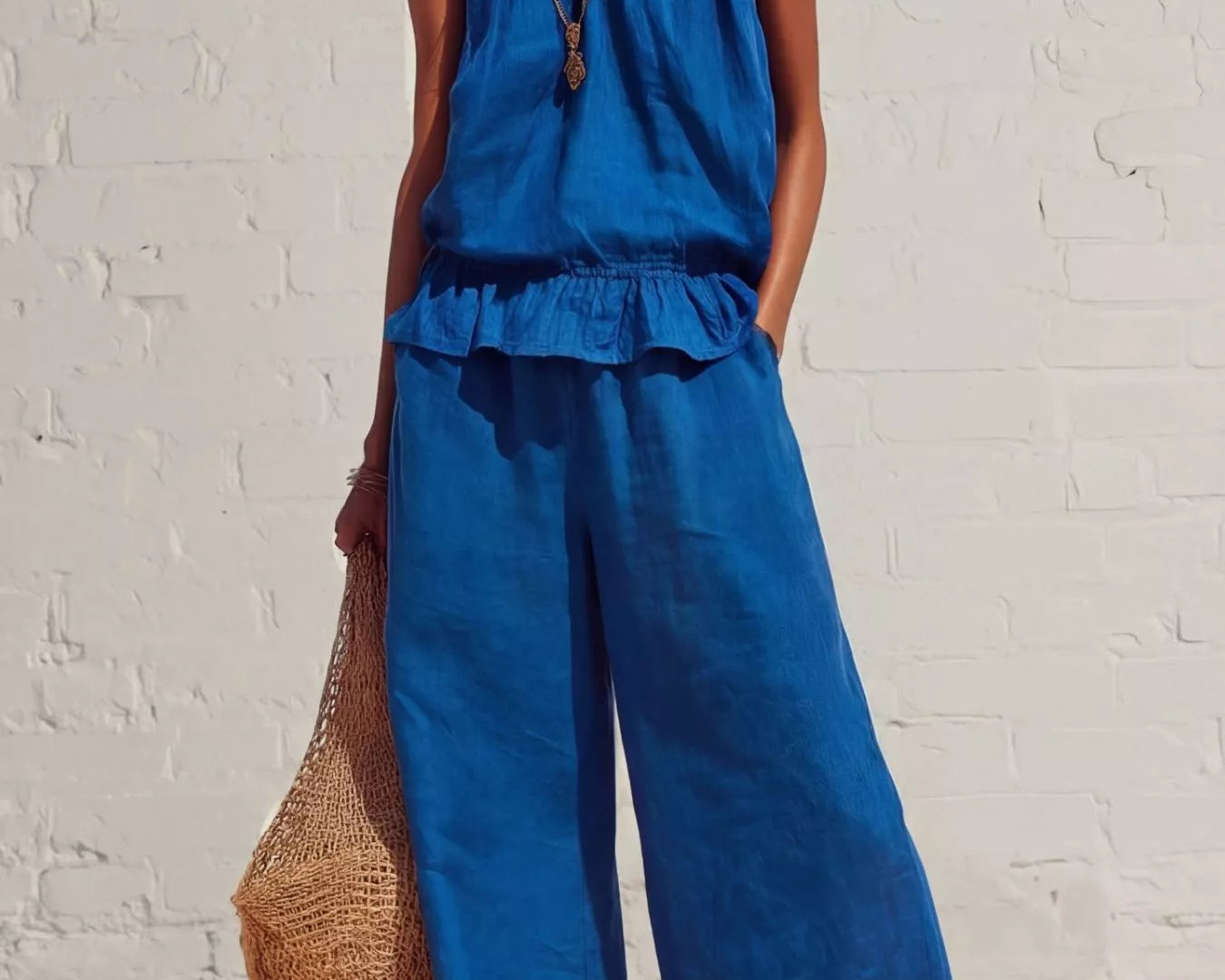 NEWYORKLISTED STYLE COLLECTION: Woman wearing a blue sleeveless top and wide-leg pants against a white brick wall.