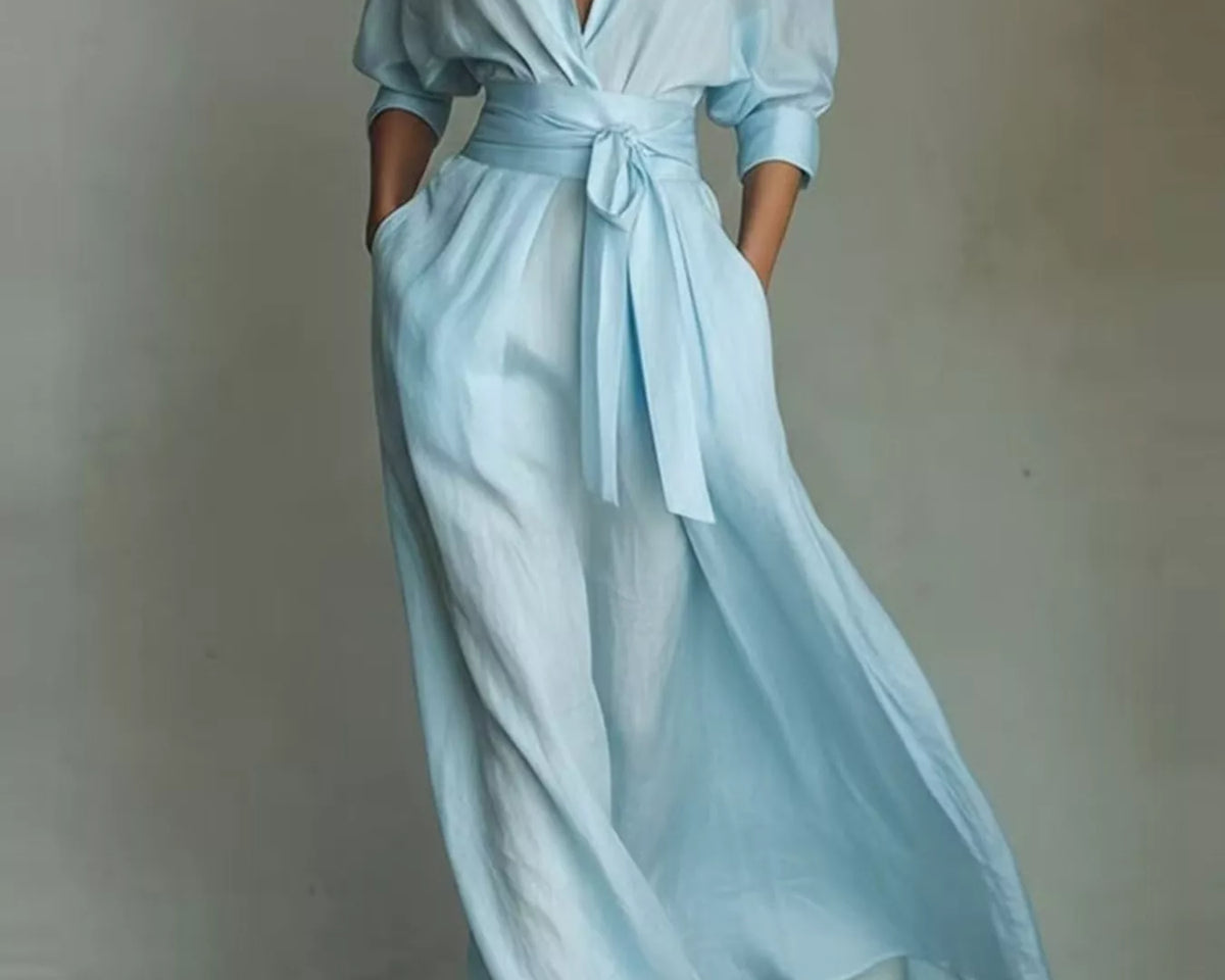 NEWYORKLISTED STYLE COLLECTION: A woman models a light blue wrap-style maxi dress with a tie waist, long sleeves rolled to the forearm, and side pockets, photographed against a neutral gray studio backdrop.