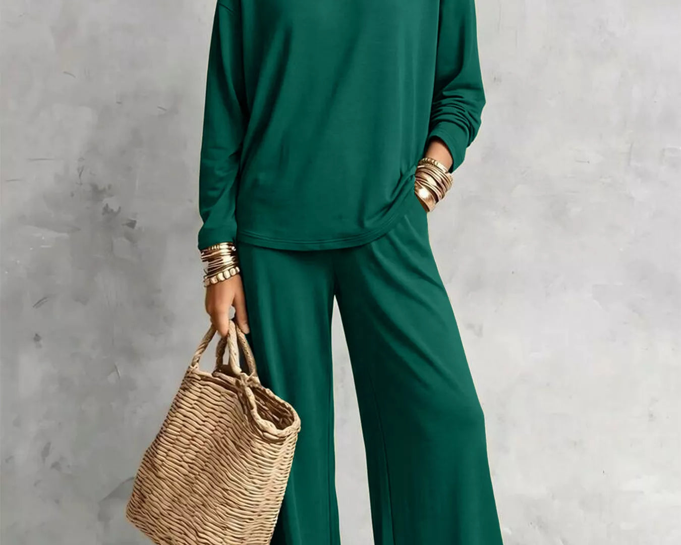 NEWYORKLISTED STYLE COLLECTION: A woman models a matching dark green long-sleeve top and wide-leg pants set, styled with layered gold necklaces, bracelets, and a woven straw tote bag against a textured gray studio backdrop.