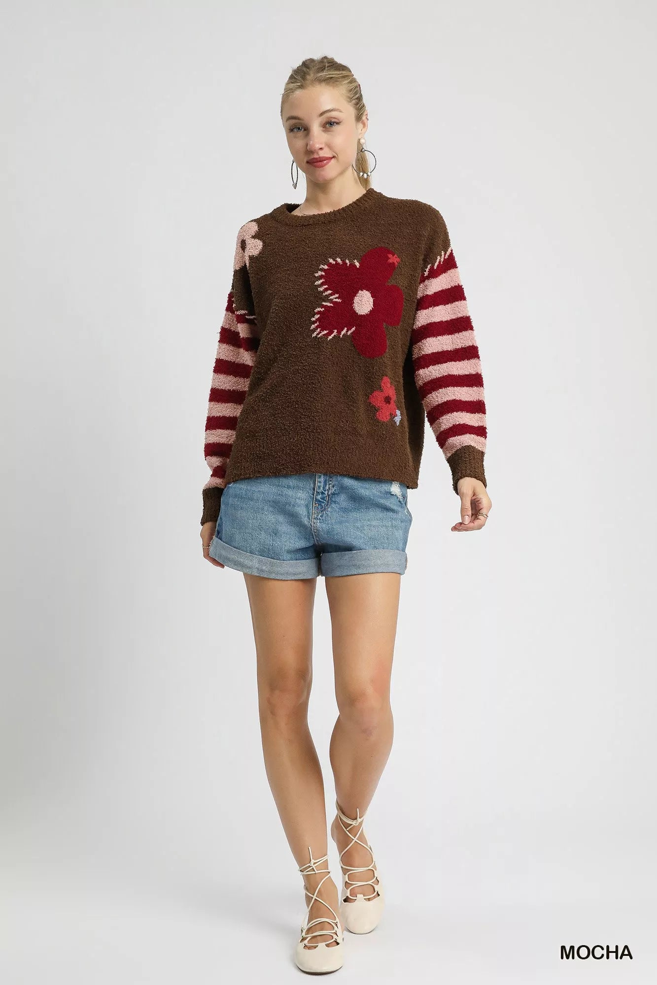 Woman wearing a brown sweater with floral designs and denim shorts on a white background: NEWYORKLISTED.COM
