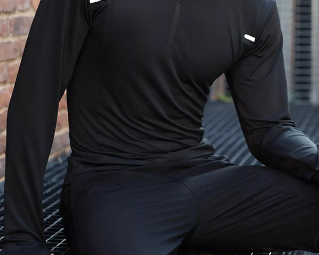 NEWYORKLISTED ATLAS MEN'S COLLECTION: Person wearing a black athletic outfit sitting on a metal bench with a cityscape background.