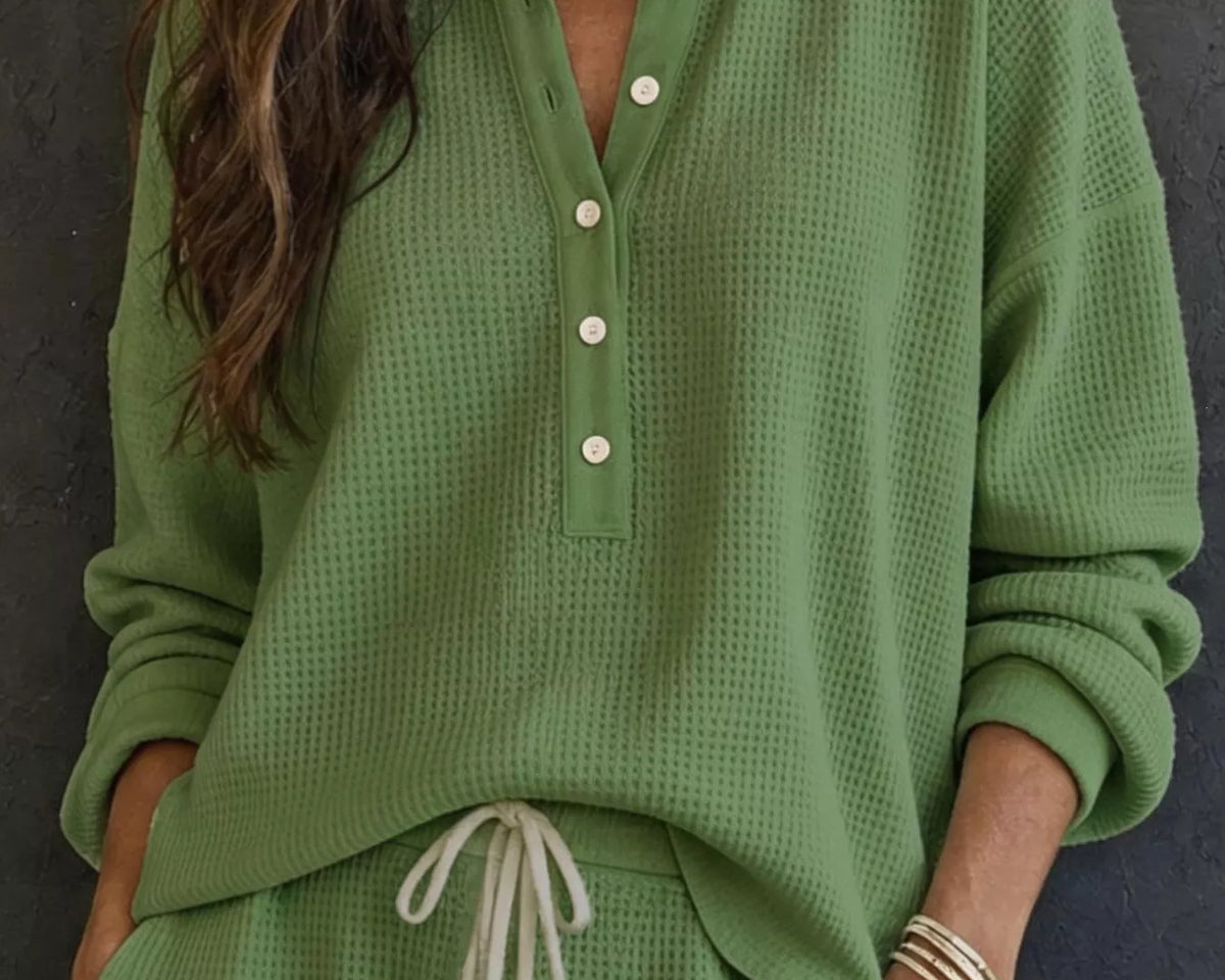 NEWYORKLISTED STYLE & COMFORT COLLECTION: A woman wearing a sage green waffle-knit long-sleeve henley top with white buttons and matching green drawstring pants, standing against a dark textured wall.