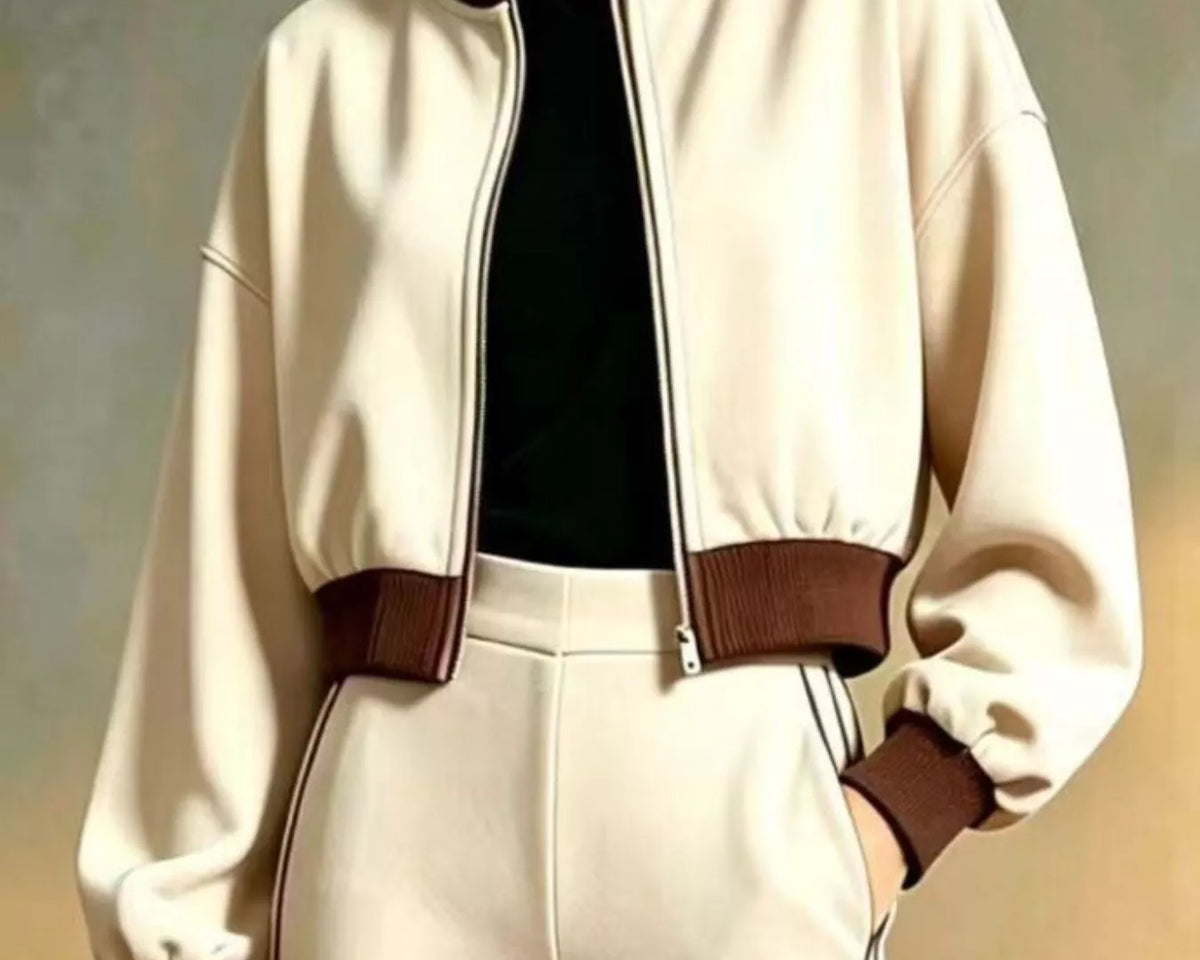 NEWYORKLISTED STYLE COLLECTION: A woman models a cream-colored bomber jacket with dark brown ribbed trim, paired with matching cream pants and a black top, set against a neutral studio background.