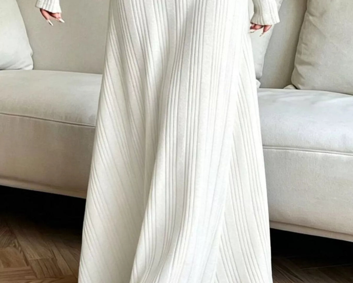 NEWYORKLISTED STYLE COLLECTION: A person wearing a long-sleeve, off-white ribbed knit maxi dress with a side slit, standing in a living room setting with a beige sofa and wooden floor.