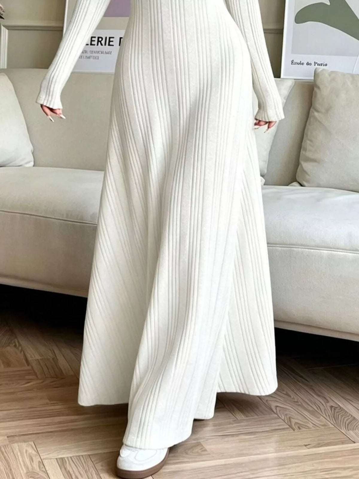 NEWYORKLISTED STYLE COLLECTION: A person wearing a long-sleeve, off-white ribbed knit maxi dress with a side slit, standing in a living room setting with a beige sofa and wooden floor.