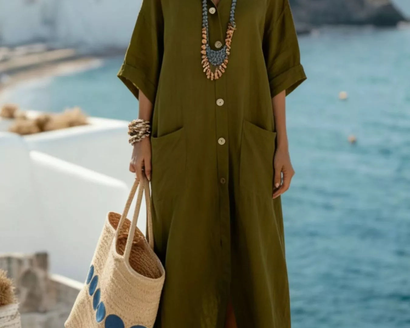 NEWYORKLISTED STYLE COLLECTION: Woman in a green dress holding a straw bag with blue patterns, standing on a balcony with ocean view.