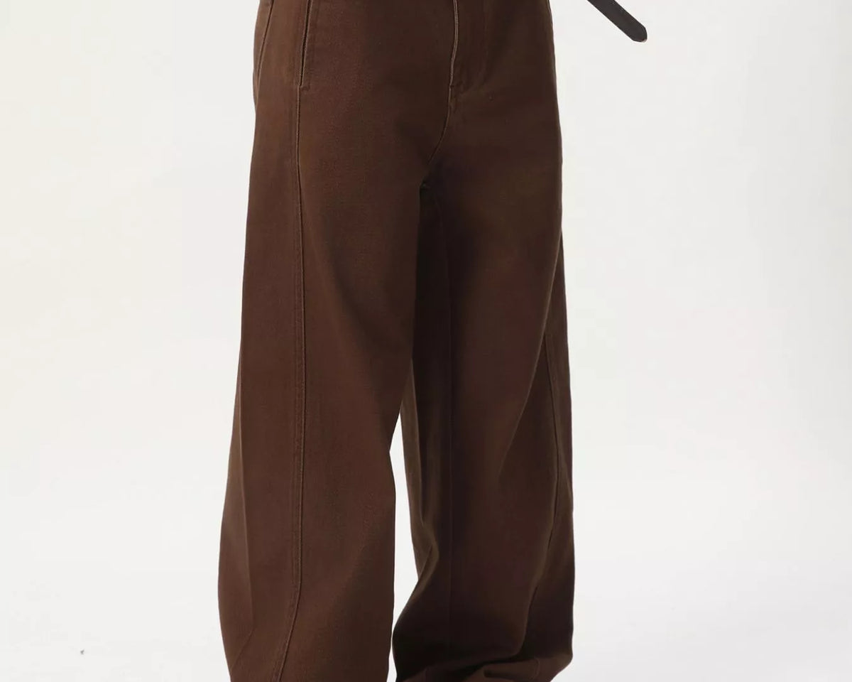 NEWYORKLISTED ATLAS MEN'S COLLECTION: A person shown from the waist down wearing loose brown wide-leg pants, a black belt, a white shirt, and tan slip-on shoes against a plain light background.