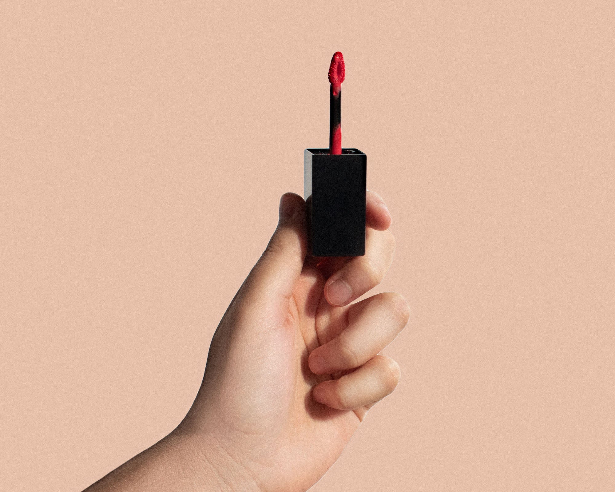 NEWYORKLISTED Cosmetic Collection: Hand holding a black lip gloss with red applicator against a beige background