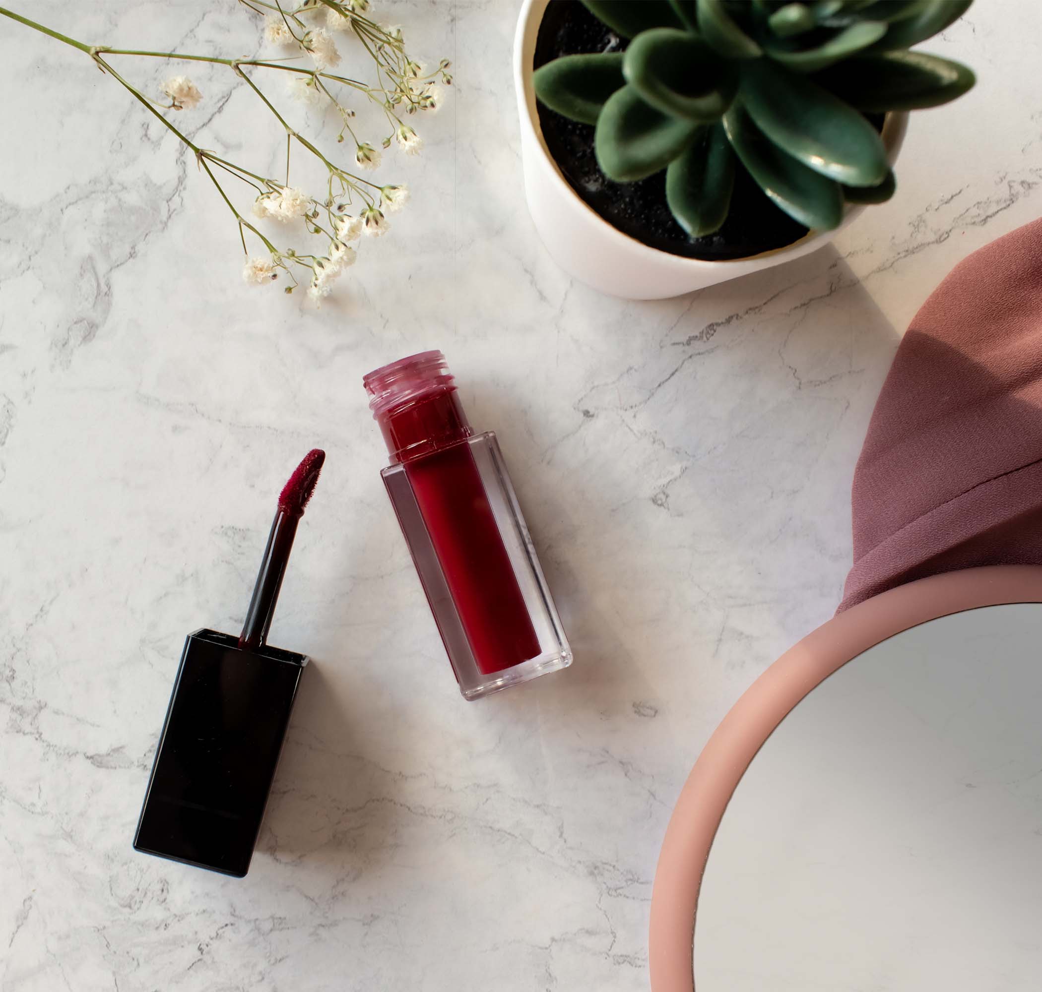 NEWYORKLISTED Cosmetic Collection: Red lip gloss and applicator on a marble surface with a plant in the background