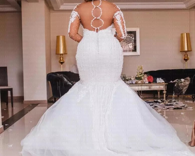 Woman in a white mermaid-style wedding dress with lace sleeves in an indoor setting.