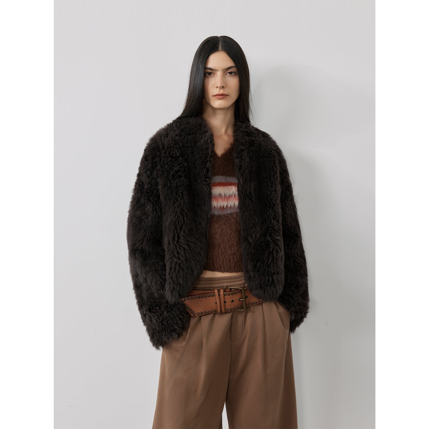 NEWYORKLISTED STYLE COLLECTION: Person wearing a dark brown fur coat over a patterned sweater and brown pants on a light gray background