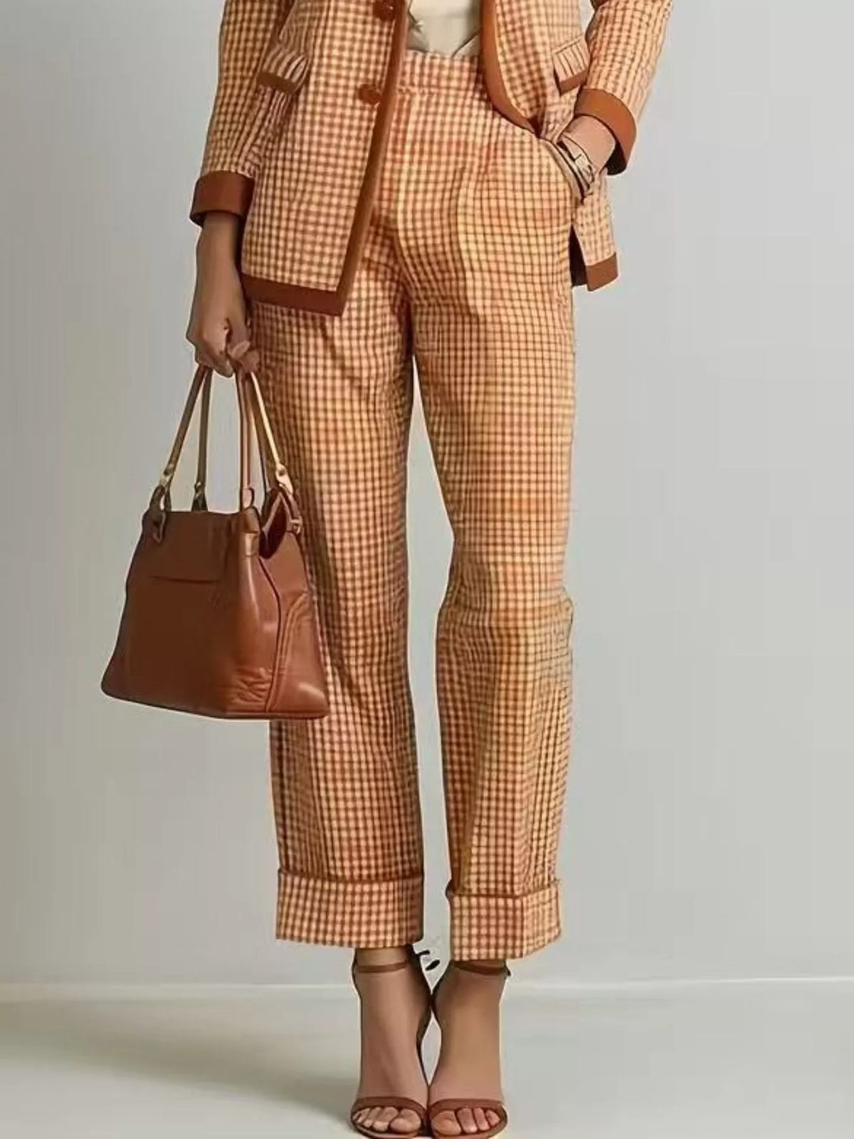NEWYORKLISTED STYLE COLLECTION: A model wearing a matching orange and beige gingham blazer and cropped trousers with brown trim, holding a brown leather handbag and wearing brown heeled sandals against a light gray studio background.