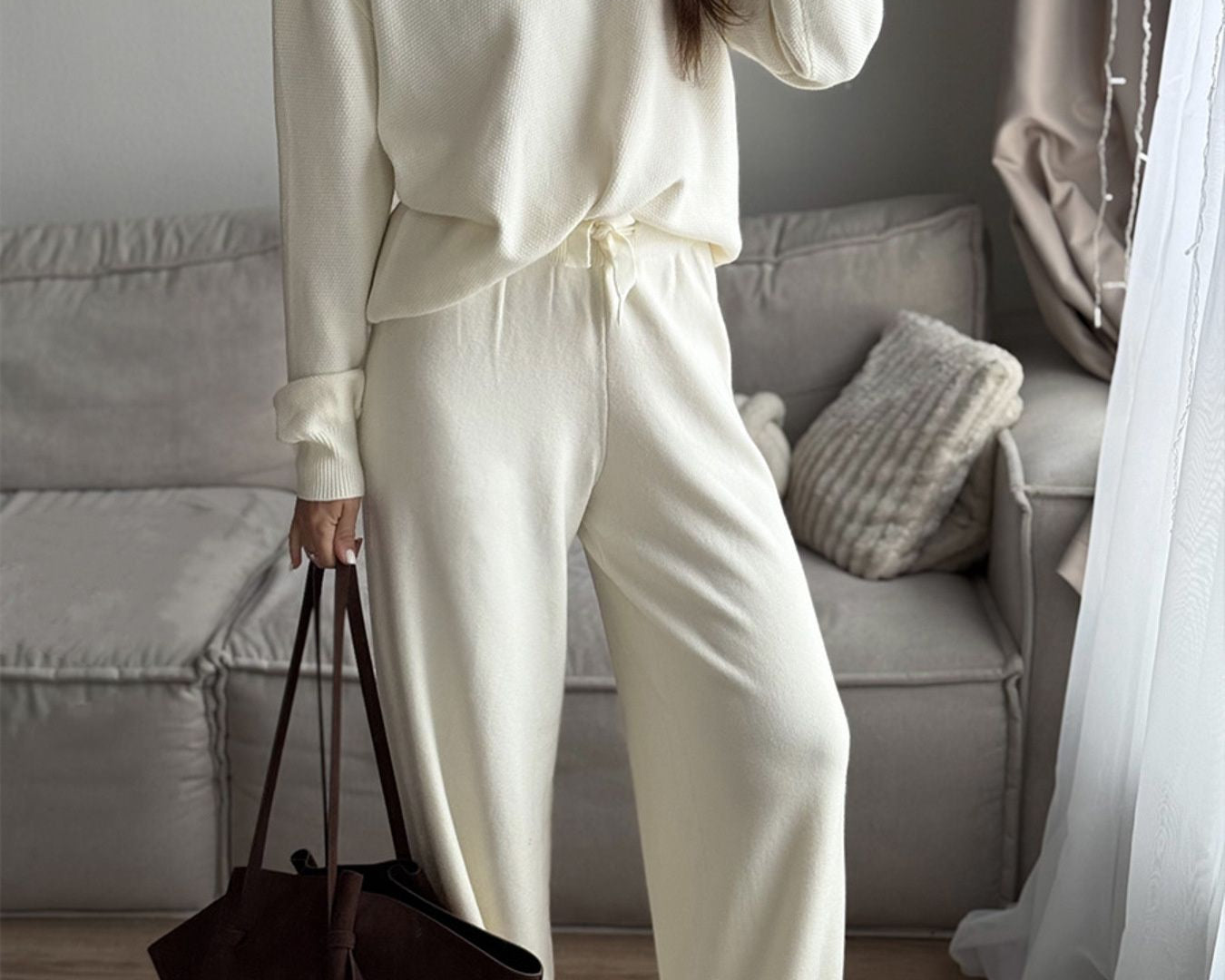 NEWYORKLISTED Comfort Collection: Woman in a white outfit holding a brown bag in a living room.