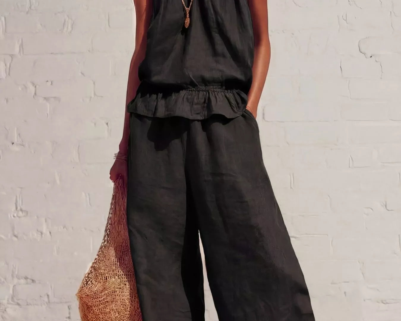 NEWYORKLISTED STYLE COLLECTION: Woman wearing a black sleeveless top and wide-leg pants against a white brick wall.