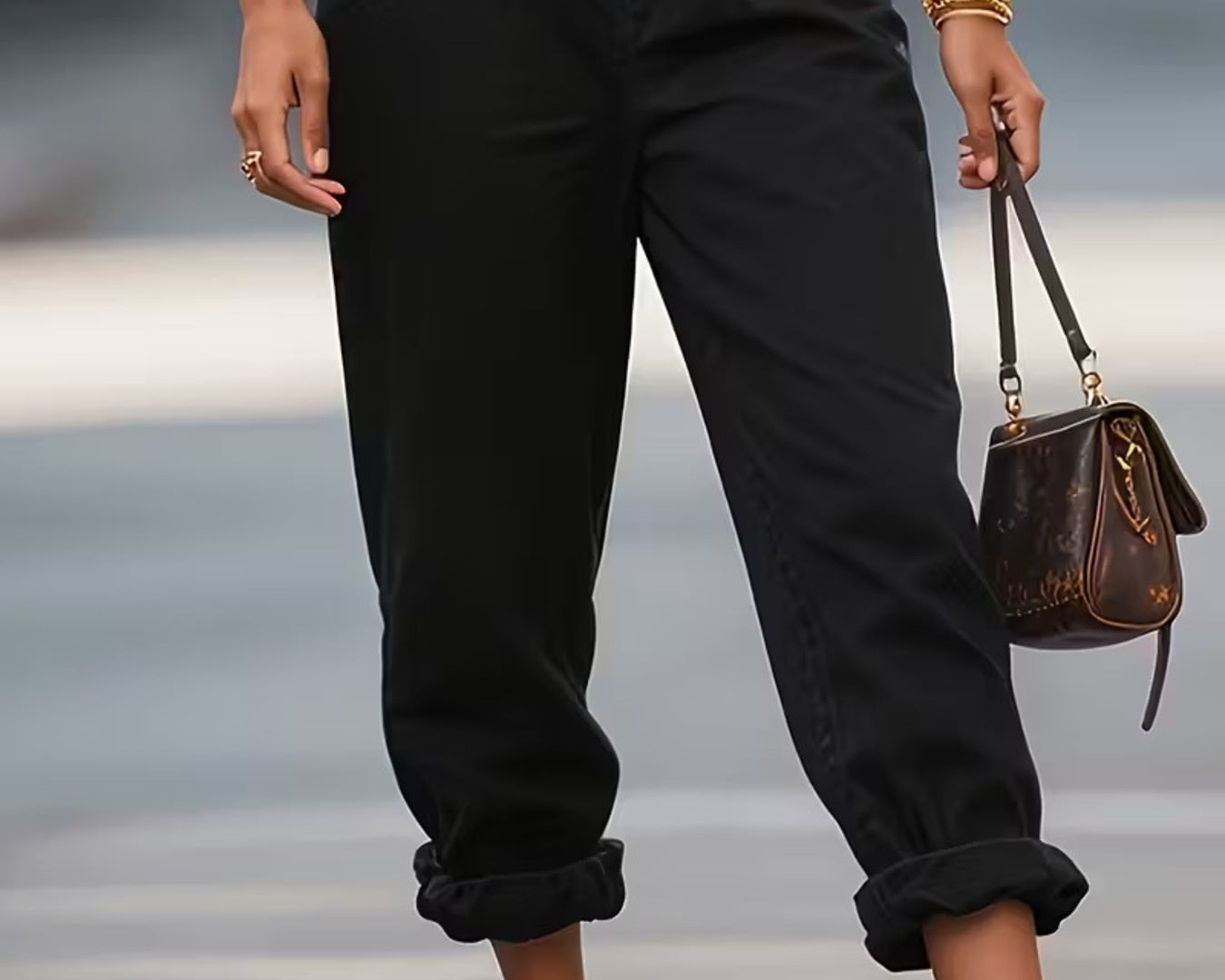 NEWYORKLISTED STYLE COLLECTION: Person wearing black pants with gold zippers, holding a brown handbag on a blurred background