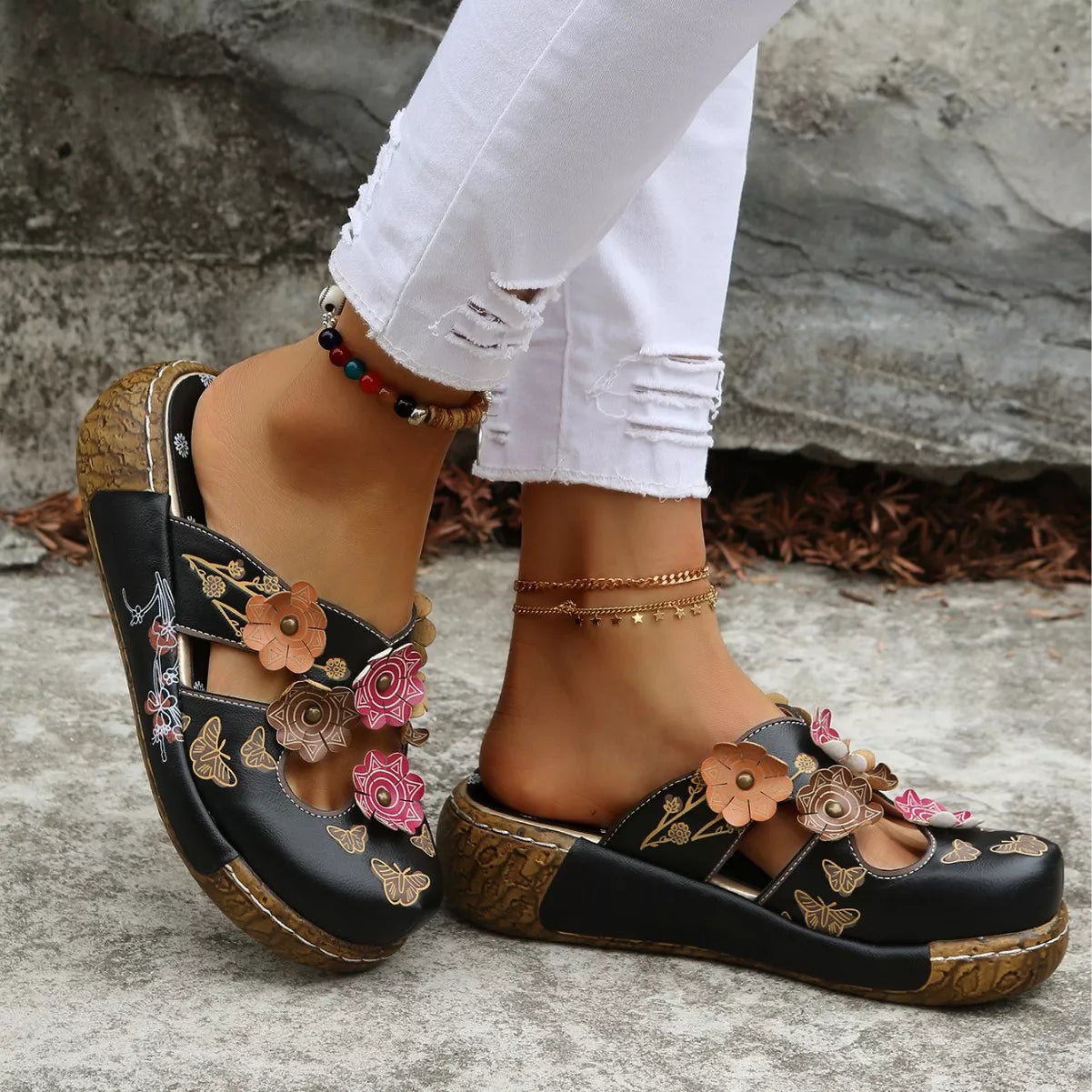 NEWYORKLISTED SHOE COLLECTION: Black sandals with floral designs worn by a person on a stone surface