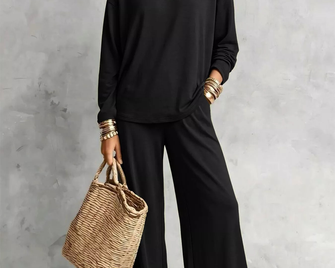 NEWYORKLISTED STYLE COLLECTION: A woman models a black long-sleeve top and matching wide-leg pants, styled with layered gold necklaces, bracelets, and a woven straw tote bag against a textured gray wall.
