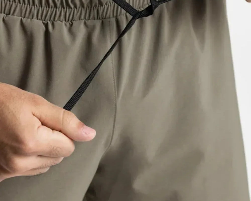 NEWYORKLISTED ATLAS MEN'S COLLECTION: Person adjusting a drawstring on olive green shorts against a white background