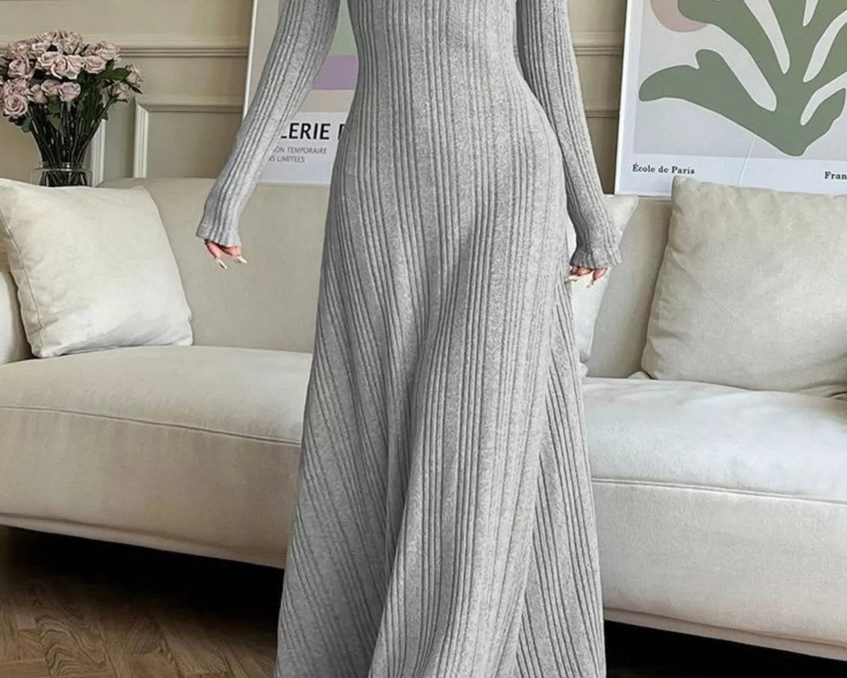 NEWYORKLISTED STYLE COLLECTION: A woman wearing a long-sleeve, ribbed knit maxi dress in light gray, standing in a living room setting with a beige sofa and framed wall art in the background.