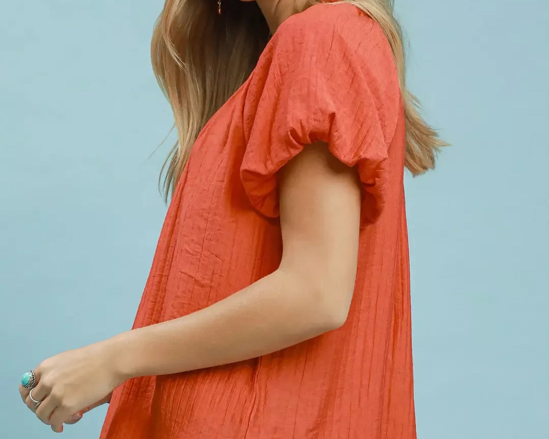 NEWYORKLISTED STYLE COLLECTION: Woman wearing a coral top with ruffled sleeves against a light blue background