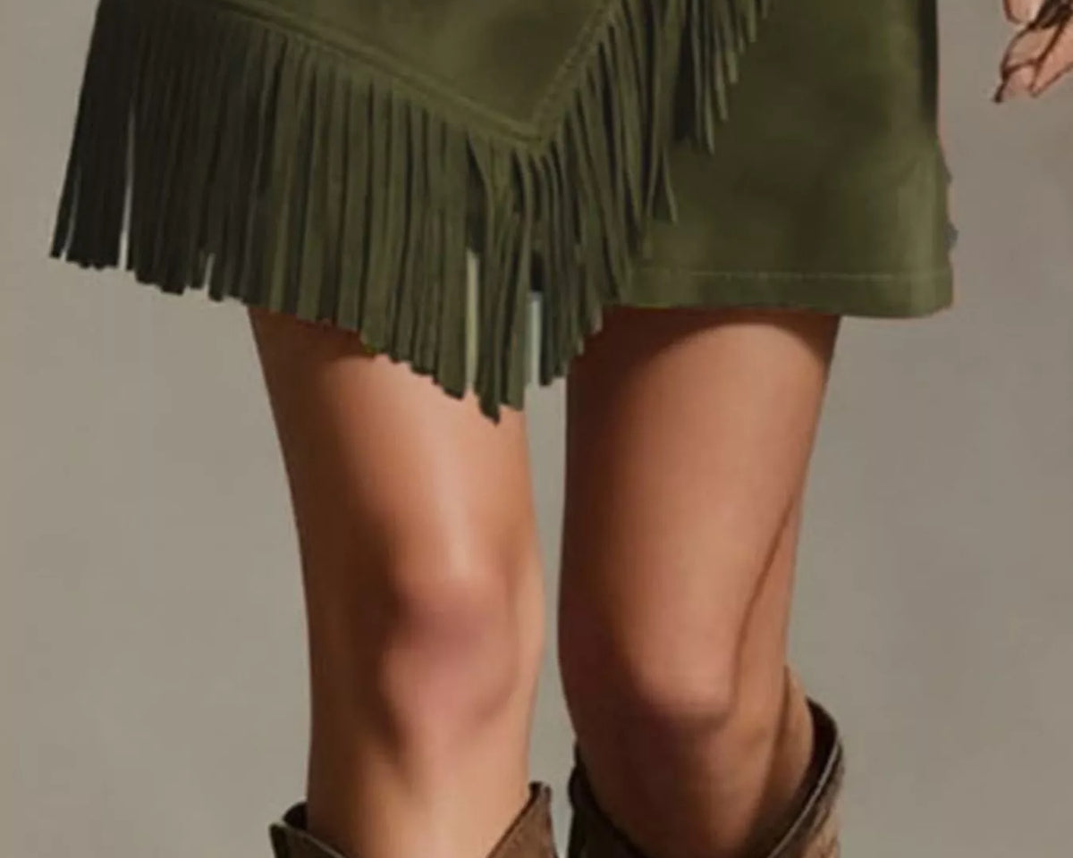 NEWYORKLISTED STYLE COLLECTION: An olive green faux-suede mini skirt with asymmetrical fringe trim, styled with brown suede cowboy boots against a neutral studio background.