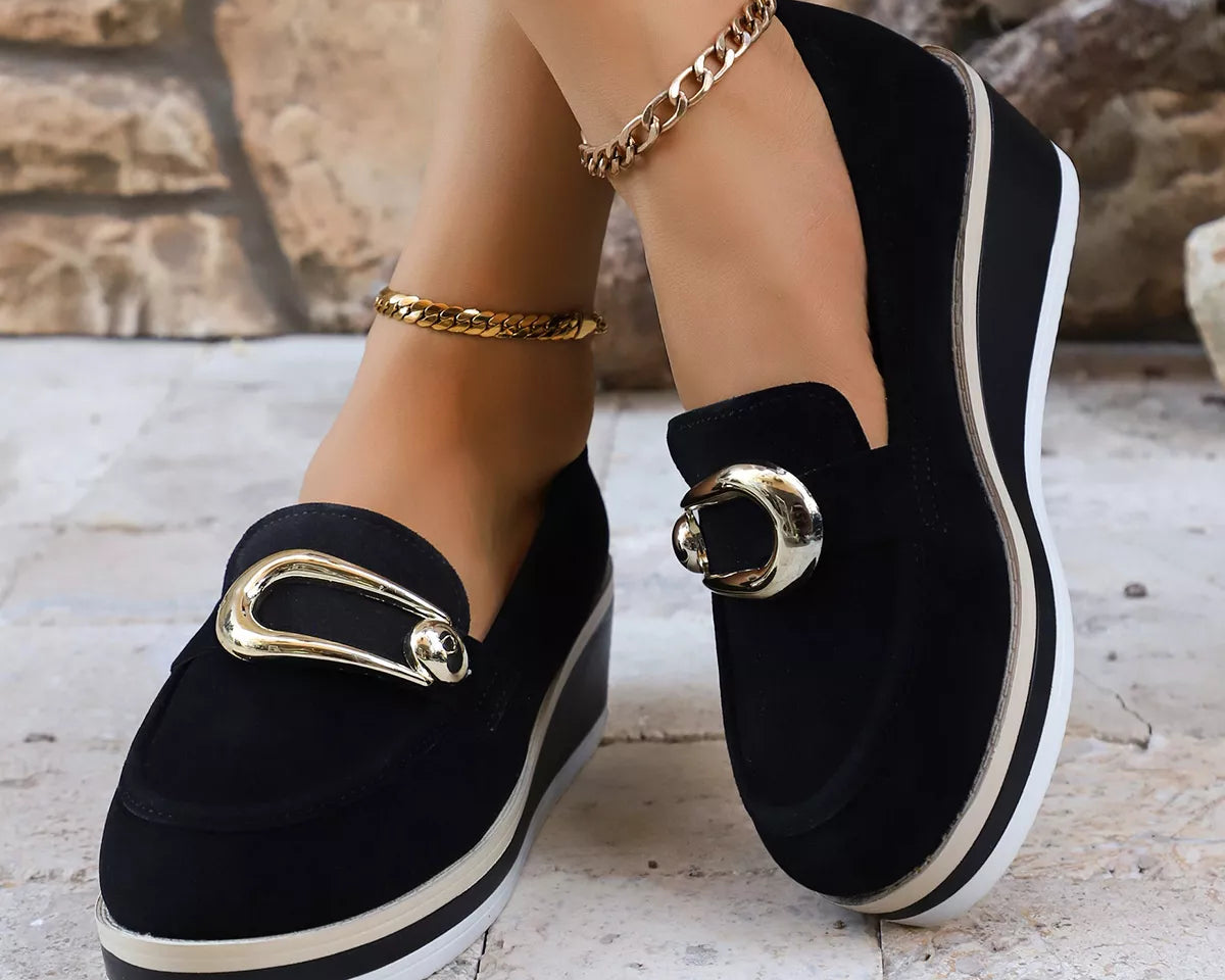 NEWYORKLISTED SHOE COLLECTION: Black loafers with gold buckles worn by a person against a stone wall background