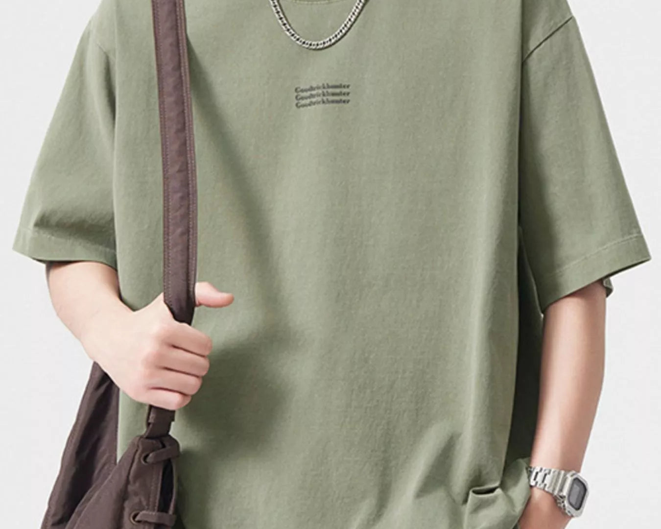 NEWYORKLISTED ATLAS MEN's COLLECTION: Person wearing a green t-shirt with a subtle logo, holding a brown bag on a plain background